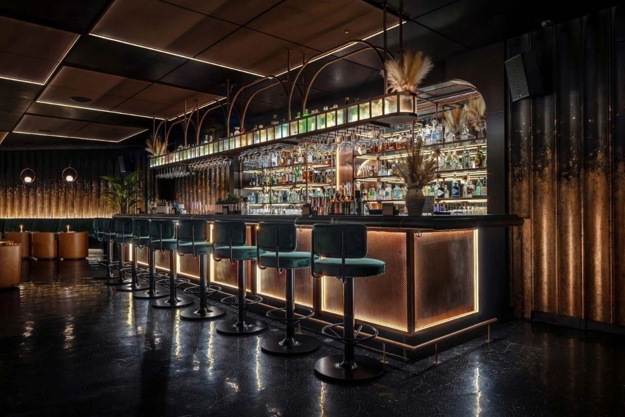The backlit bar at HUSH seen from an angle, with velvet bar stools and pampas grass on the shelves